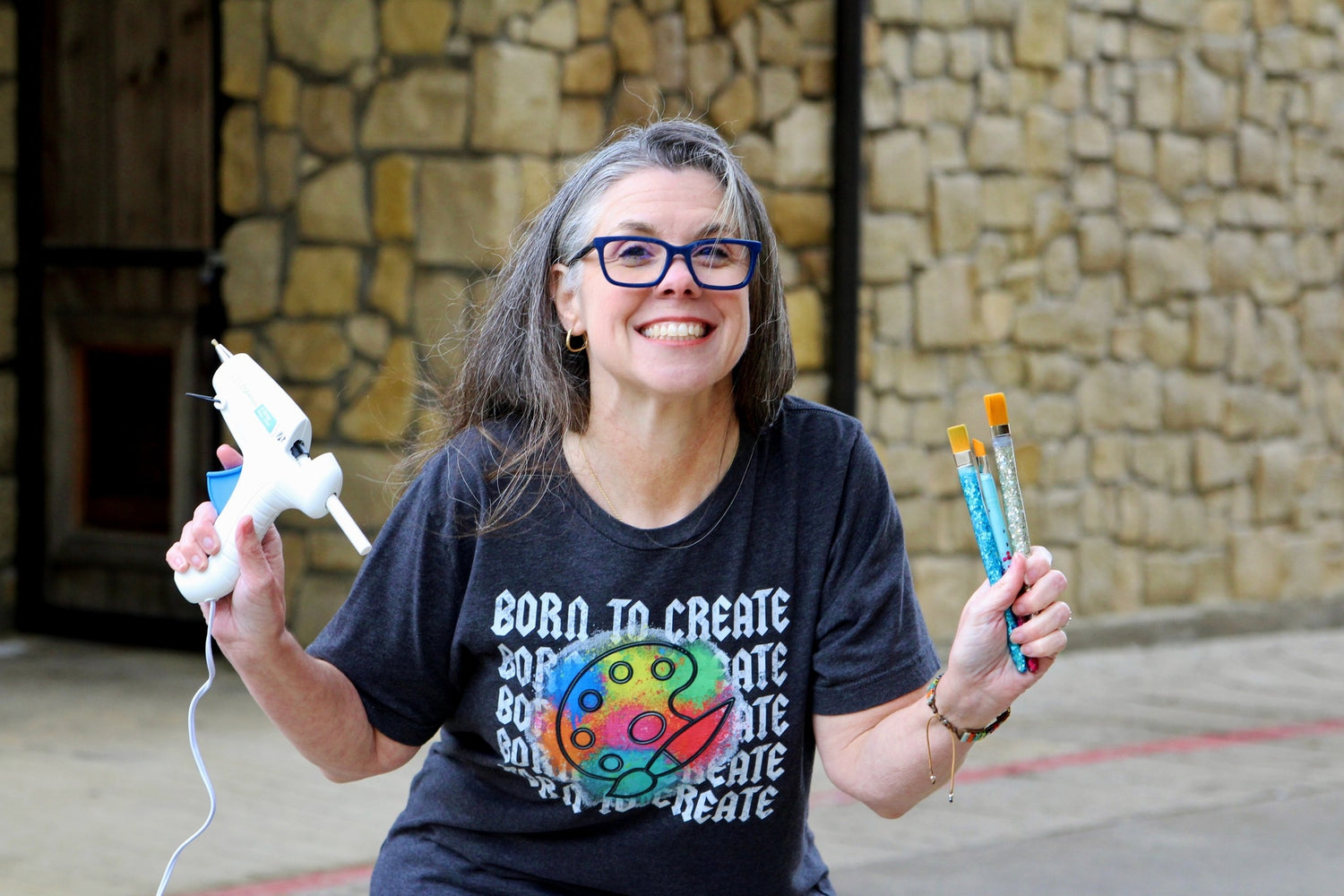 Amie (Deep In The Arts Creations owner) poses with a smile in a black T-Shirt with born to create printed on it five times. The words have a paint pallet over it with a tie-died type of pattern using colors of blue, yellow, green, red, aqua, orange, and pink. She is holding a glue gun in her right hand and three paint brushes in her left. 